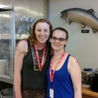 Two women stand smiling into the camera, with a mounted fish on the wall behind them.