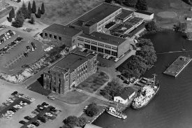 An aerial black and white photo of a group of buildings located near the water, which has a dock and boats moored up.