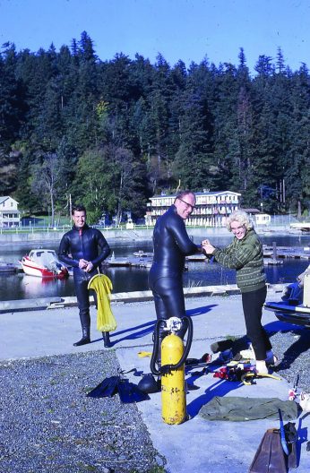 Two people in diving gear and another person stand on docks by the water.