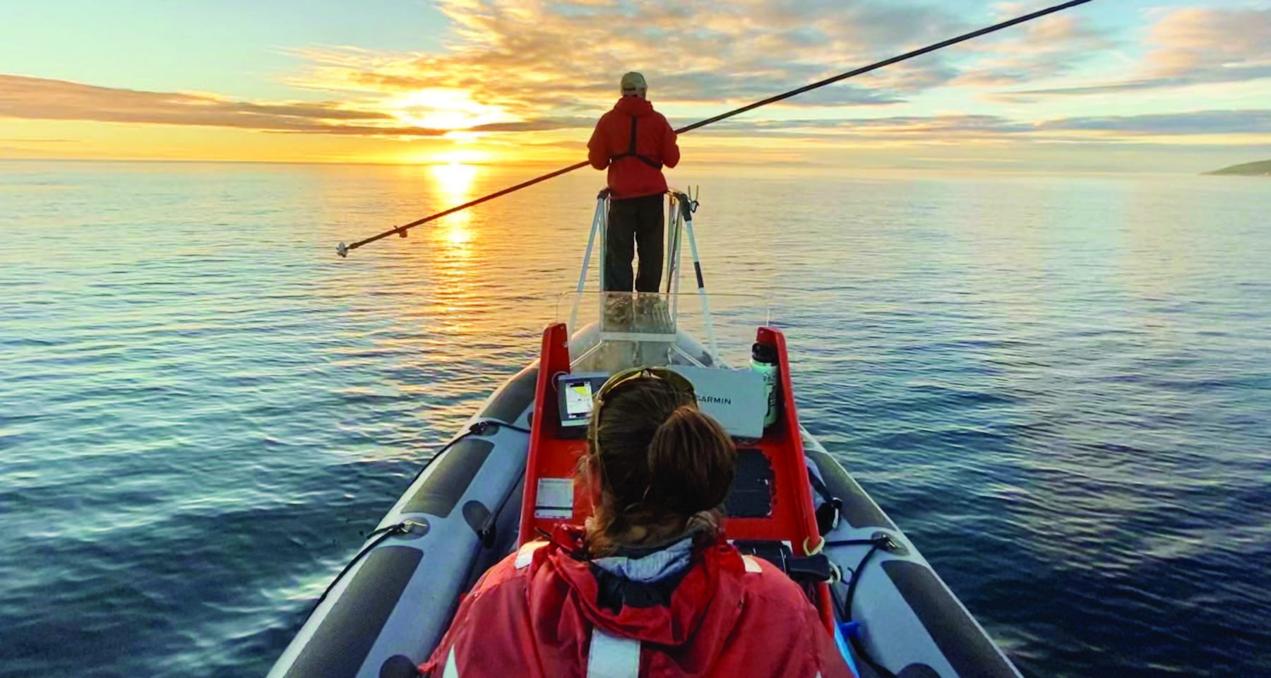 Two people, one sitting and one standing, pictured on a boat on calm water with a sunset in the distance. Both wear red coats, and the person standing at the front of the boat holds a long fishing pole horizontally.