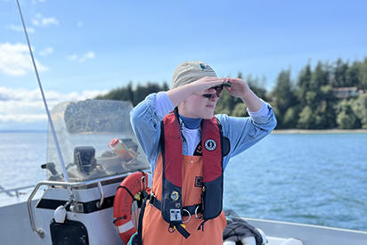 A person on a boat wearing orange waders and a red life jacket, is pictured staring into the distance. Calm water and blue skies with white clouds can be seen in the background.