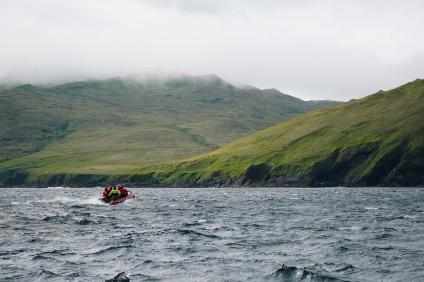 A small boat holding a group of people heads over choppy water towards land. The land has tall cliffs, green grass, and low misty cloud.