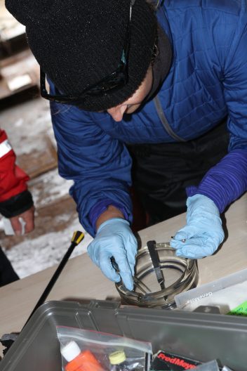Kim bends over a table, wearing blue gloves as she prepares to pick up a sample from a clear glass jar holding tweezers.