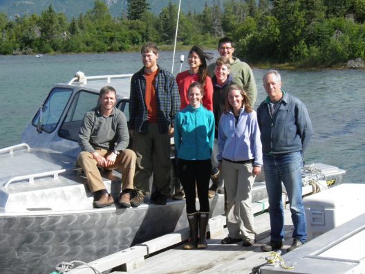 The AERA class of 2012 stands on a boat and a dock, smiling in the camera, located on a lake with greenery in the background.