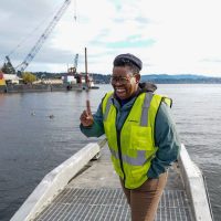Ashley Townes, wearing a high-vis vest, smiles into the camera, while standing on a dock with a lake in the background.