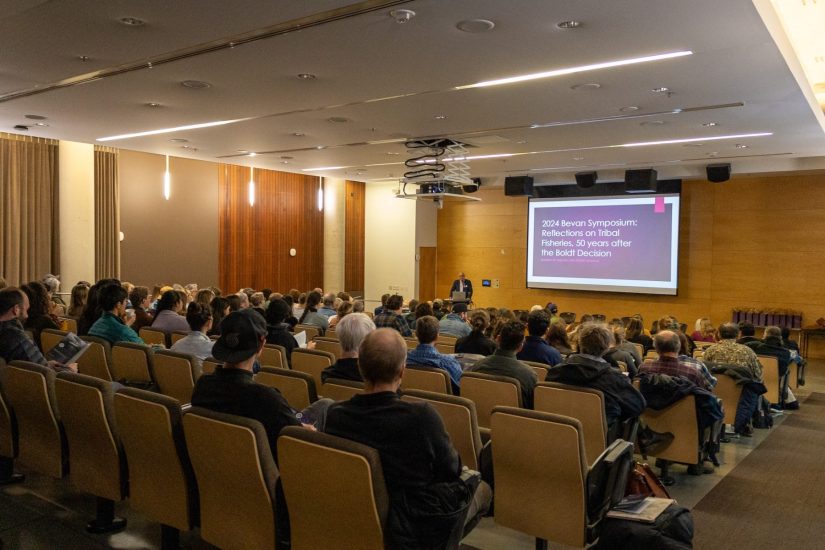 A crowded room faces a presentation screen, with one person at the lectern.