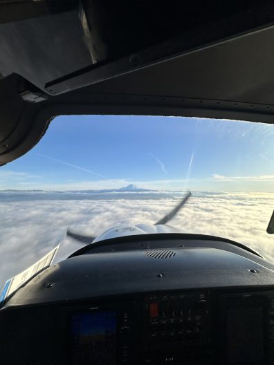 An image from the cockpit of a small plane, flying over white clouds with a mountain in the distance.