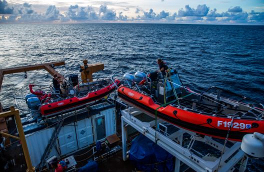 Two, smaller red boats pictured on the deck of a larger ship, sailing on the ocean.