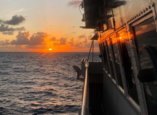 A view of an orange sunrise from the side of a boat on the ocean.
