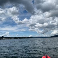 The tip of a red kayak is pictured on a large lake, surrounded by wooded area. Big fluffy white clouds are visible in the sky.