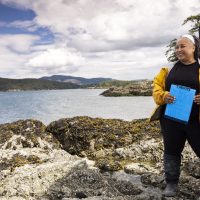 Chris Mantegna, wearing a yellow rain jacket and holding a blue clipboard, stands on a rocky outcropping with a body of water and another island behind her. A blue sky with white clouds is above.