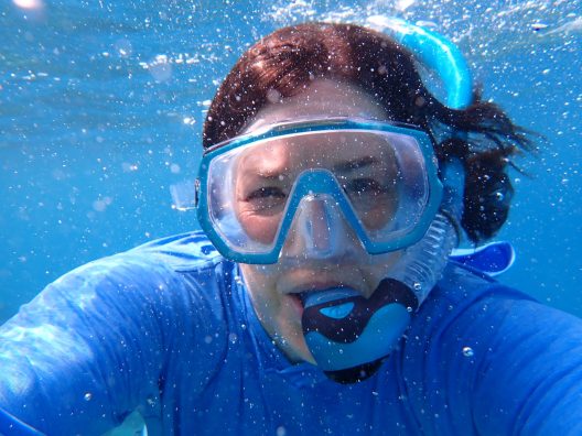 A selfie of person wearing a blue tshirt, underwater with a snorkel mask on.