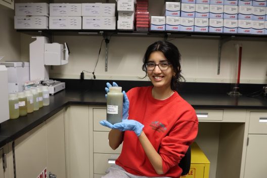 A woman, smiling and wearing a red sweatshirt, blue lab gloves and glasses, sits in a lab holding a container that holds green-grey liquid. The container has a lid and a white label with black writing on.