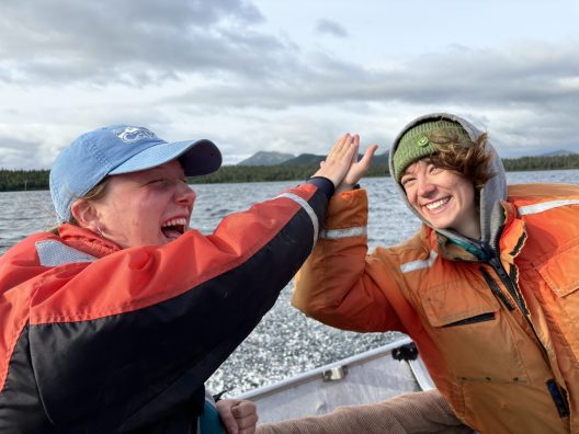 Two people high-five on a boat with choppy water, low hills and a cloudy sky behind them. The person on the left is smiling with eyes closed, a blue baseball cap on, and wearing a black and red jacket. The person on the right smiles into the camera wearing a green beanie, a grey sweatshirt with the hood up, and an orange coat.