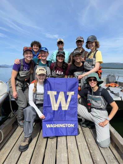 A group of people holding a purple flag with the large, gold W for University of Washington, stand and kneel on a dock smiling into the camera.