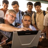 A group of people sit and stand around a laptop screen.