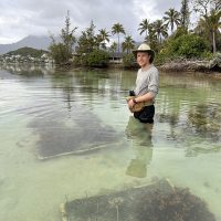 Callum stands hip-deep in water, with corals visible on the shallow seafloor in front of him. In the background, the coastline with palm trees is visible.