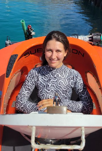 Juliette sits in an orange submersible, with blue water visible in the background.