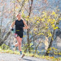 Kristina is pictured running on a trail with trees and a river in the background.