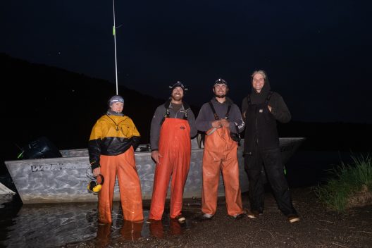 A group of people stand at night in front of a boat wearing bright orange wading gear.