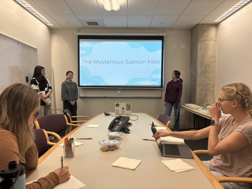 Three students stand at the front of the room to give a presentation, while two people sit at the table to watch.