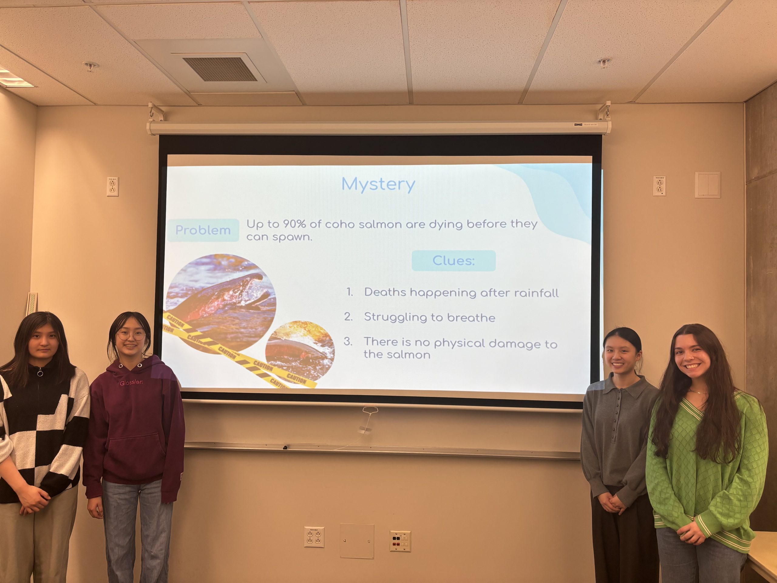 Four students (three high school and one graduate) stand in front of a presentation screen.