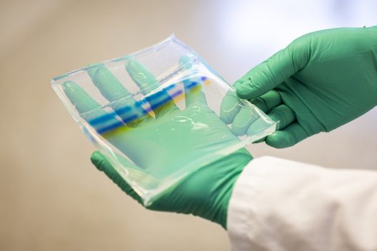Hands wearing green gloves hold a square clear dish with ribbons of color.