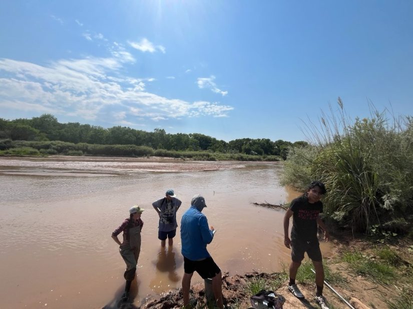 Four people stand in, and on the banks of, the Rio Grande.