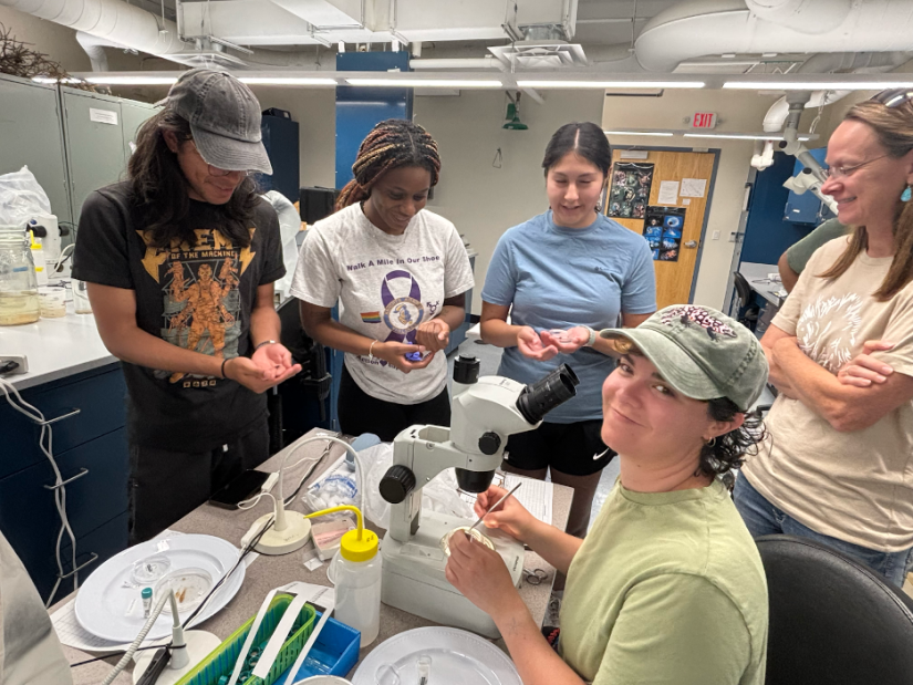 A group of people in a lab sit and stand around a microscope.