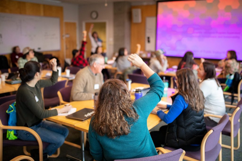 A group of people sitting at various tables in a classroom, some with their hands raised.